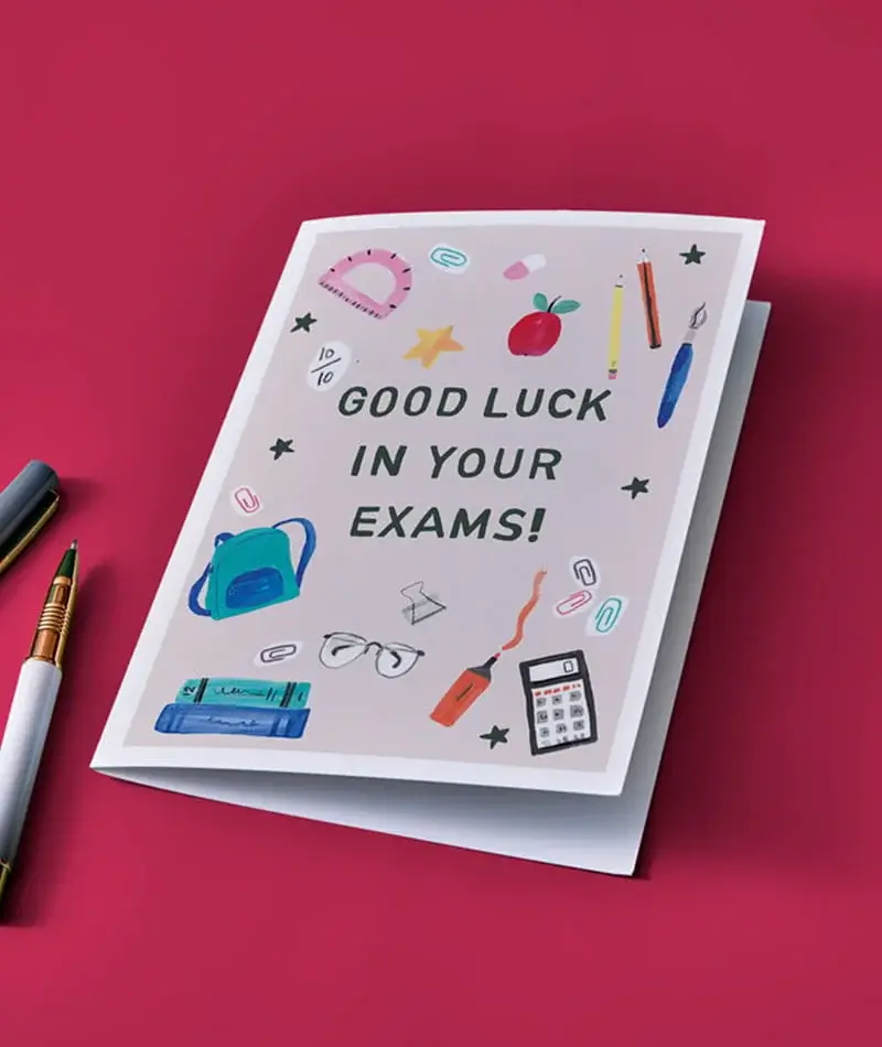 A good luck in your exams greeting card decorated with colourful school-themed illustrations, placed on a bright pink background beside two pens. A good luck in your exams greeting card decorated with colourful school-themed illustrations, placed on a bright pink background beside two pens.
